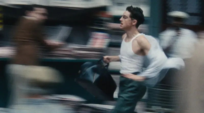 Crítica de 'Marty Supreme': Retrato de una obsesión. 1 Fotograma en movimiento de la película 'Marty Supreme'. Timothée Chalamet, caracterizado como el jugador de ping-pong Marty Reisman, corre velozmente en una calle concurrida. Viste una camiseta de tirantes blanca, pantalones de vestir oscuros y una camisa azul claro que ondea al viento por la velocidad. Lleva un maletín de cuero oscuro en la mano. La imagen presenta un marcado efecto de desenfoque de movimiento (motion blur) que resalta la urgencia de la escena.
