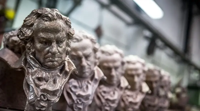Fila de estatuillas de los Premios Goya alineadas en perspectiva, con el primer busto de bronce nítidamente enfocado mostrando el rostro de Francisco de Goya, y el resto difuminándose progresivamente hacia el fondo.
