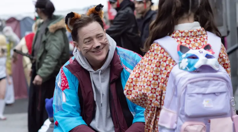 Fotograma de la película 'Rental Family'. Primer plano medio de Brendan Fraser sonriendo con ternura mientras se agacha para estar a la altura de una niña (cuya espalda vemos). Él lleva una diadema con orejas de zorro y bigotes pintados en la cara, imitando la máscara de zorro que cuelga de la mochila lila de la niña. El fondo muestra una multitud borrosa en lo que parece un festival cultural.