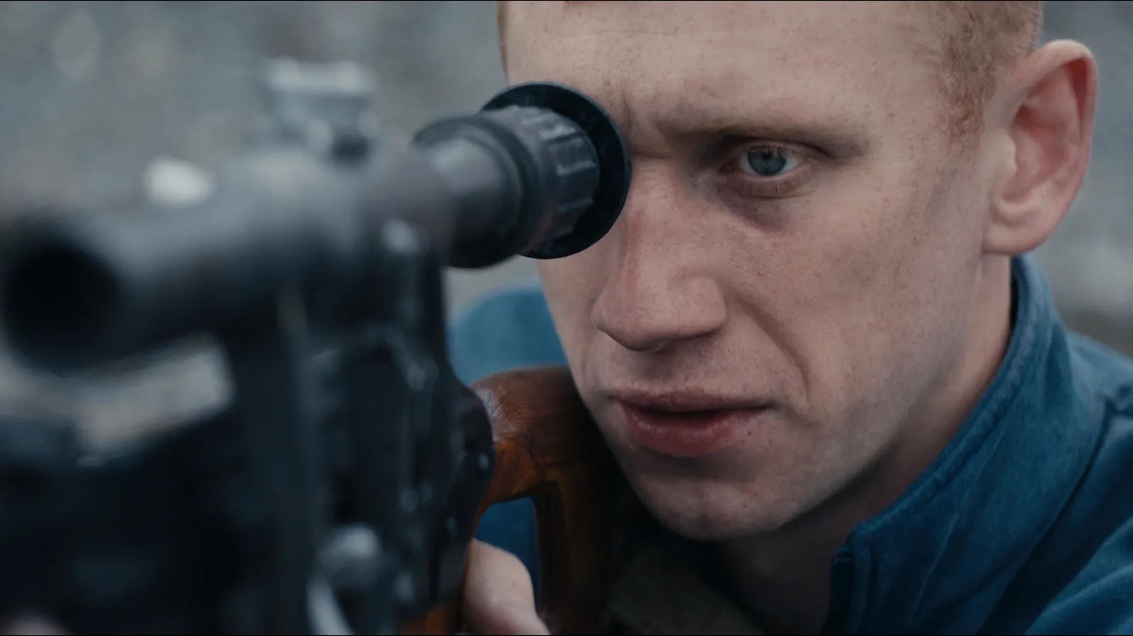 Primer plano lateral extremo de un joven con cabello corto rojizo y ojos azules mirando intensamente a través de la mira telescópica de un rifle de francotirador con culata de madera.