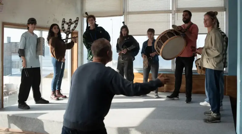 Fotograma de la película 'Rondallas'. En una sala amplia y luminosa con grandes ventanales al fondo, un grupo de siete jóvenes músicos posa con instrumentos tradicionales gallegos. Se observa una gaita, panderetas, un bombo y un charrasco. En primer plano, de espaldas al espectador y con los brazos abiertos en gesto de dirección, se encuentra un hombre con un jersey azul. Los músicos, vestidos con ropa informal contemporánea, miran hacia el director con una mezcla de atención y expectación. La luz natural inunda la estancia, creando un ambiente de ensayo auténtico y comunitario.