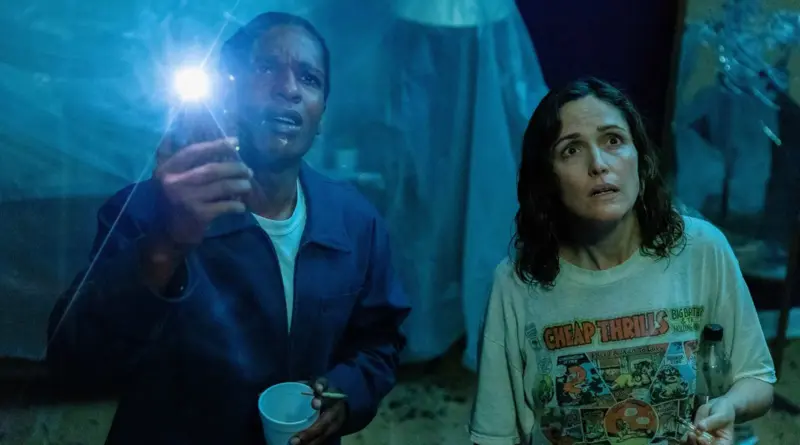 Fotograma de la película 'Si pudiera te daría una patada' en un interior oscuro bañado por luz azul y linternas. El rapero A$AP Rocky, vistiendo una chaqueta de trabajo azul, ilumina hacia arriba con una linterna y expresión de sorpresa. A su lado, Rose Byrne, con una camiseta gráfica que dice "Cheap Thrills", mira en la misma dirección con preocupación. El fondo parece estar cubierto de plásticos.