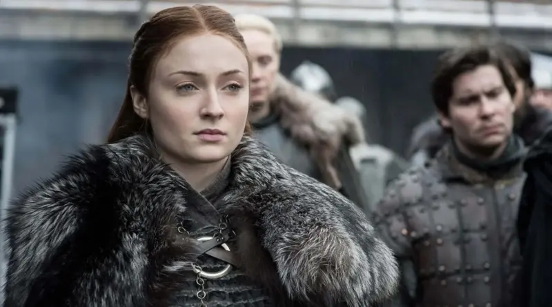 Sophie Turner caracterizada como Sansa Stark con expresión seria y regia, vistiendo una capa de piel gris oscura y el broche circular de los Stark, con Brienne de Tarth y Podrick Payne desenfocados al fondo.