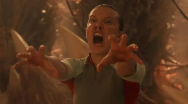 Primer plano de Eleven (Millie Bobby Brown) con el rostro desencajado por el esfuerzo, gritando y extendiendo ambas manos hacia adelante mientras sangra por la nariz en un entorno oscuro y polvoriento en 'Stranger Things'.