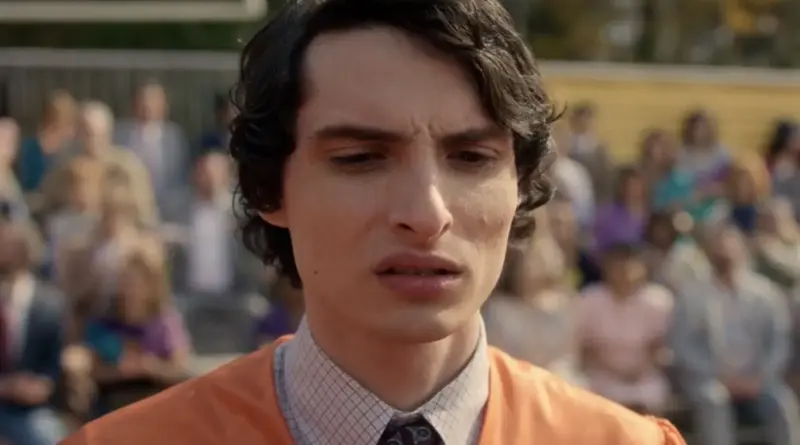 Primer plano de Mike Wheeler con ceño fruncido, vistiendo una túnica de graduación naranja sobre una camisa de cuadros durante la ceremonia de cierre de curso en el final de 'Stranger Things'.