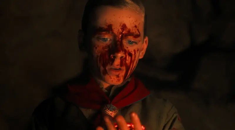Un niño, Henry Creel de 'Stranger Things', con el rostro cubierto de sangre y un pañuelo rojo al cuello observa con fascinación un objeto brillante de color rojo intenso que sostiene entre sus manos en la penumbra.