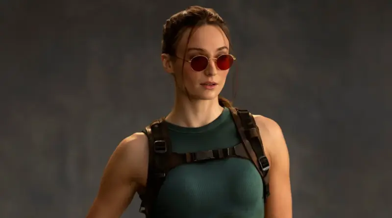 Primer plano medio de Sophie Turner caracterizada como Lara Croft en la serie ?Tomb Raider', vistiendo una camiseta de tirantes verde azulado, un arnés de cuero marrón y unas gafas de sol redondas con cristales tintados de rojo, mirando hacia su izquierda sobre un fondo gris neutro.