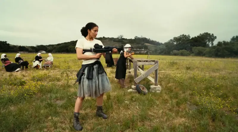 Fotograma de la película 'Una batalla tras otra'. La actriz Chase Infinity está de pie en un campo de hierba con flores amarillas, sosteniendo un rifle de asalto pesado o ametralladora ligera con ambas manos. Viste una camiseta blanca, falda gris, una chaqueta de cuero atada a la cintura y botas de combate. Al fondo, un grupo de mujeres con pañuelos blancos en la cabeza (con aspecto de monjas o lugareñas) están sentadas en sillas de jardín observando, mientras otra persona dispara desde un banco de tiro.