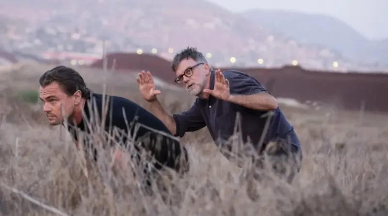 Fotografía del rodaje de 'Una batalla tras otra', la nueva película de Paul Thomas Anderson. El director Paul Thomas Anderson (derecha), con gafas y camisa gris, hace gestos con las manos dirigiendo la escena. A su lado, Leonardo DiCaprio (izquierda) aparece agachado en sigilo entre hierba seca, vistiendo una camiseta negra. Mira intensamente hacia el horizonte. Al fondo, desenfocado, se distingue una estructura larga que parece un muro fronterizo o valla bajo una luz de atardecer.