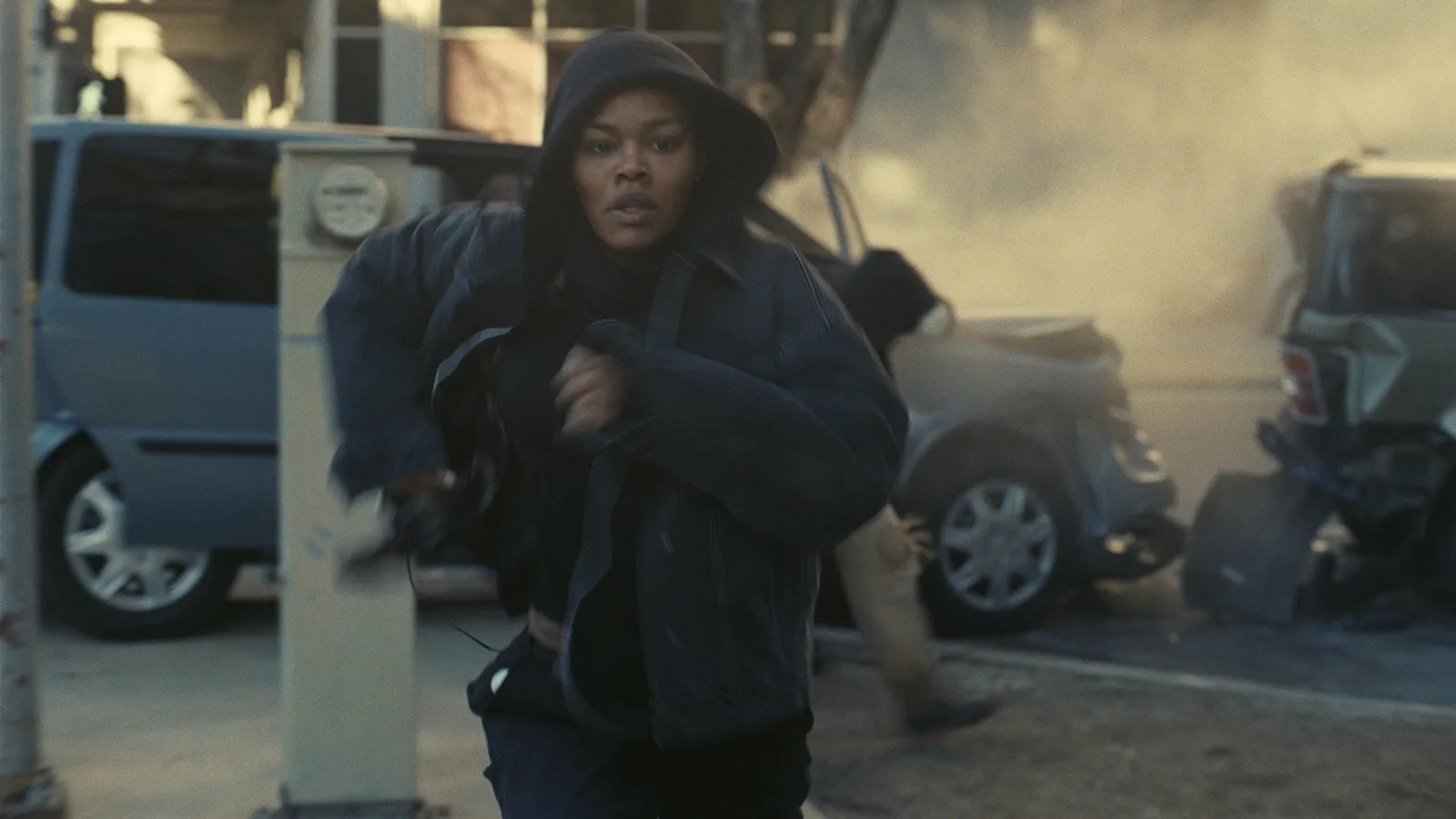 Fotograma de acción de 'Una batalla tras otra' con la actriz Teyana Taylor corriendo frontalmente hacia la cámara con expresión de urgencia y determinación. Viste una chaqueta vaquera oscura con capucha y detrás de ella se ve una calle sumida en el caos, con humo denso, escombros y coches accidentados.