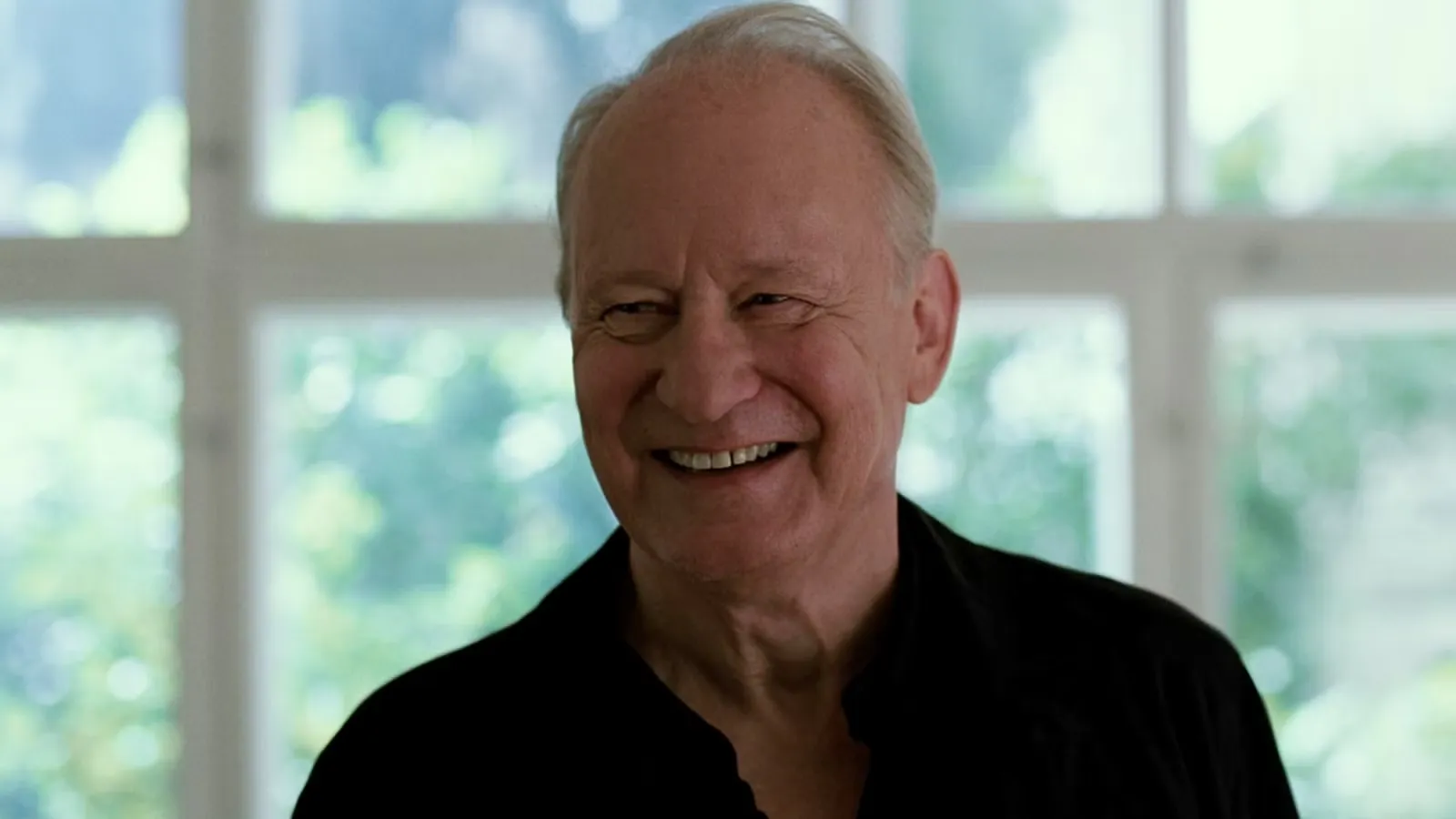 Primer plano medio del actor Stellan Skarsgård sonriendo ampliamente y mirando hacia un lado en 'Valor Sentimental'. Viste una camisa negra sencilla. El fondo está desenfocado, mostrando una ventana grande con marcos blancos y vegetación verde brillante en el exterior.