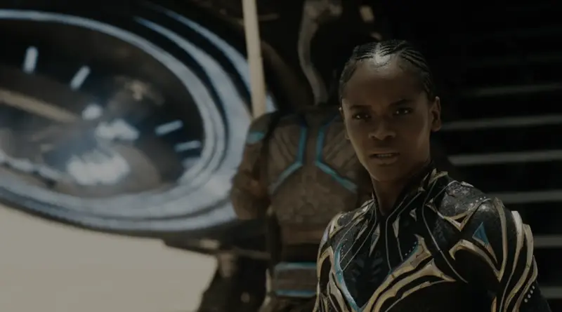 Letitia Wright como Shuri en 'Vengadores: Doomsday' con un nuevo traje de Pantera Negra negro y dorado con detalles azules, mirando fijamente a la cámara con determinación, delante de una gran turbina tecnológica y un guerrero wakandiano desenfocado.