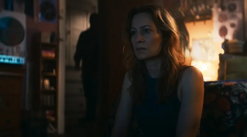 Fotograma De 'La Maldición De Shelby Oaks'. Plano Medio De La Protagonista Sentada En El Borde De Una Cama O Silla En Una Habitación En Penumbra. Mira Hacia El Vacío Con Terror Paralizante. Al Fondo, Desenfocada En El Marco De La Puerta Abierta, Se Recorta Una Silueta Masculina Oscura Que La Observa.