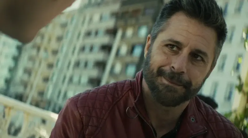 Fotograma De 'Cada Día Nace Un Listo'. Primer Plano De Hugo Silva Como Toni Lomas. Viste Su Característica Chaqueta De Cuero Roja Acolchada Y Mira A Alguien Fuera De Cámara Con Una Sonrisa Pícara Y Confianza En Su Rostro, Con Un Edificio Urbano De Fondo.