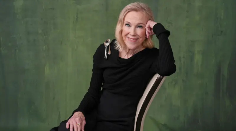 Retrato de estudio de Catherine O'Hara sentada en una silla de perfil, apoyando la cara en su mano y sonriendo con dulzura a cámara, vistiendo un top negro sobre un fondo verde texturizado.