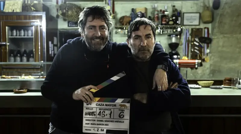Imagen detrás de las cámaras de 'Caza mayor' en el interior de un bar rústico. El director Daniel Sánchez Arévalo (izquierda) sonríe mientras sostiene una claqueta de producción frente al actor Antonio de la Torre (derecha), quien aparece caracterizado con una herida en la frente y expresión seria.