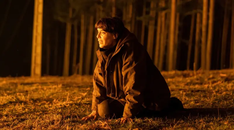 Fotograma de 'Cortafuego'. Belén Cuesta está arrodillada en el suelo de un bosque por la noche. Todo el fondo está iluminado por un resplandor anaranjado intenso y se ven troncos de árboles silueteados por el fuego. Ella mira hacia un lado con terror y agotamiento.