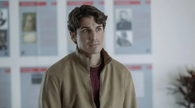 Fotograma De La Serie Day One. Primer Plano De Álex González (Ulises) Vistiendo Una Chaqueta Beige Sobre Un Jersey Granate. Mira Hacia Un Lado Con Una Expresión Melancólica Y Reflexiva En Un Espacio Amplio Y Luminoso. Al Fondo Se Ven Paneles Con Rostros Difuminados.