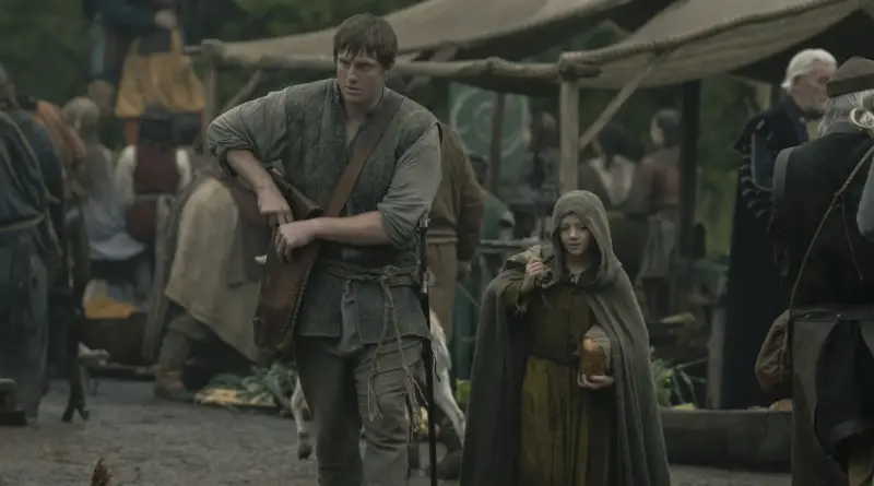 Fotograma De 'El Caballero De Los Siete Reinos'. Ser Duncan El Alto (Peter Claffey) Y Egg (Dexter Sol Ansell) Caminan Juntos Por Un Mercado Medieval Al Aire Libre; Duncan Lleva Una Gran Bolsa De Cuero Al Hombro Mientras El Niño, Encapuchado, Carga Con Un Trozo De Pan.