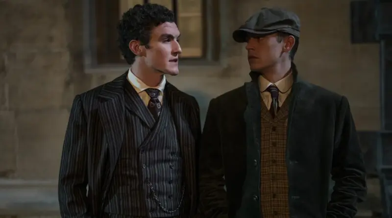 Fotograma de 'El joven Sherlock'. James Moriarty (Dónal Finn) y Sherlock Holmes (Hero Fiennes Tiffin) hablando de noche frente a un edificio de piedra. Moriarty viste un traje de rayas diplomáticas con chaleco, mientras Sherlock lleva una gorra y abrigo marrón de cuadros.