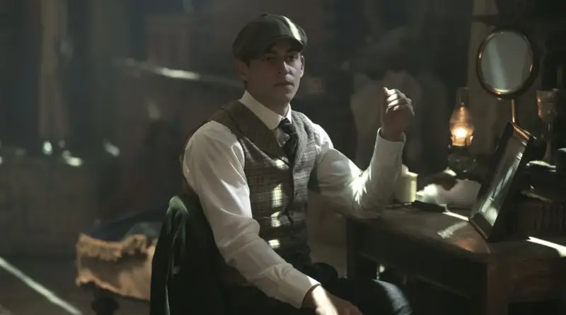 Fotograma de 'El joven Sherlock'. Sherlock Holmes sentado en un escritorio oscuro iluminado por una lámpara de aceite. Lleva puesto un chaleco de cuadros y mira con intensidad hacia adelante, con un marco de fotos y equipo de investigación sobre la mesa.