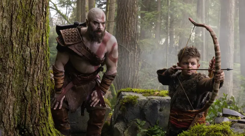 Fotograma De La Serie 'God Of War'. En Un Bosque Frondoso Con Árboles Cubiertos De Musgo, Ryan Hurst Caracterizado Como Kratos (Piel Pálida, Barba Espesa Y Tatuaje Rojo En El Torso) Observa Con Atención. A Su Lado, El Niño Callum Vinson (Atreus) Tensa Un Arco De Madera Apuntando Hacia La Derecha, Vestido Con Pieles Nórdicas.