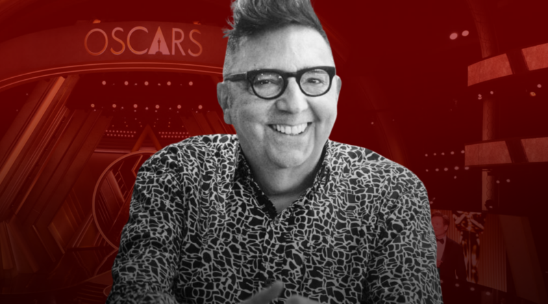 Composición gráfica. En primer plano, un recorte en blanco y negro de Guillermo de Mulder riendo, con una camisa estampada de leopardo o formas abstractas y sus características gafas de pasta. El fondo está teñido de rojo oscuro y muestra el escenario del Dolby Theatre con los arcos concéntricos y el logotipo "OSCARS" iluminado.