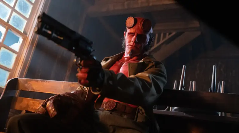 Fotograma de 'Hellboy: El Hombre Retorcido'. Plano contrapicado de Hellboy sentado en un banco de madera en un interior rústico. Apunta directamente hacia el frente con su icónico revólver de gran calibre (el "Samaritan"). La iluminación lateral crea un fuerte claroscuro sobre su rostro rojo y su gabardina, dándole un aspecto serio y amenazante.