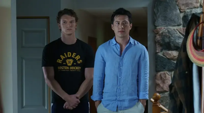 Fotograma de 'Más que rivales' ('Heated Rivalry'). Plano medio de Ilya (izquierda) y Shane (derecha) de pie en el interior de una casa. Ilya viste una camiseta negra con el logo "Raiders Boston Hockey" y mira al frente con seriedad. Shane lleva una camisa azul desabrochada en el cuello y manos en los bolsillos, con expresión neutra.