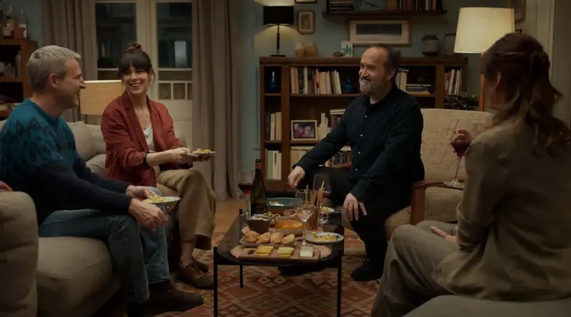 Fotograma de 'Sentimental'. Escena de interior en un salón acogedor donde cuatro personas están sentadas alrededor de una mesa baja con vino y aperitivos. A la izquierda, Javier Cámara y Belén Cuesta ríen, mientras Alberto San Juan sonríe al otro lado.