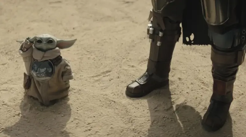 Fotograma De 'Star Wars: The Mandalorian And Grogu'. Plano Detalle A Nivel Del Suelo. Se Ven Las Botas Blindadas Del Mandaloriano A La Derecha. A Su Lado, El Pequeño Grogu Está De Pie En La Arena Vistiendo Su Túnica Beige Y Un Medallón Con El Símbolo Del Mitosaurio. Grogu Levanta Su Mano Derecha Usando La Fuerza.