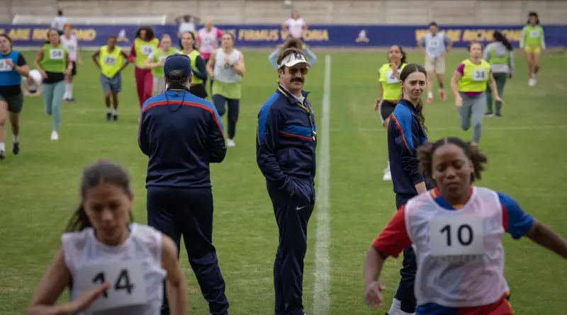 Fotograma de la temporada 4 de 'Ted Lasso'. Plano general de un entrenamiento en el campo de césped. Ted Lasso se gira mirando hacia atrás con las manos en los bolsillos. Detrás de él, un grupo numeroso de jugadoras de fútbol femeninas corre haciendo ejercicios de calentamiento bajo la supervisión de otros técnicos.
