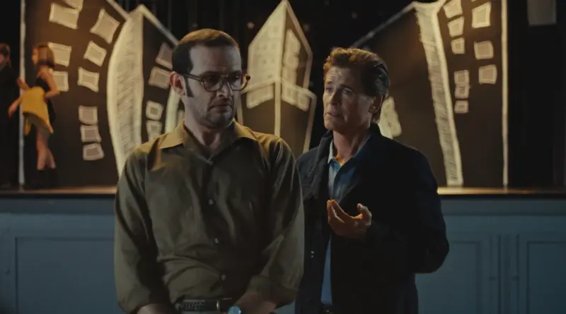 Fotograma de 'The Musical'. Plano medio en un escenario teatral con iluminación tenue. A la izquierda, Will Brill (con gafas y camisa marrón) mira hacia abajo con expresión de preocupación o vergüenza. A la derecha, Rob Lowe le habla gesticulando con la mano, con una expresión de consejo o reproche suave. El fondo muestra decorados de cartón pintado que simulan edificios torcidos y una niña actuando al fondo.