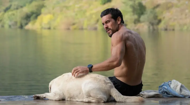 Fotograma de 'Zeta'. Mario Casas, sin camiseta y mostrando musculatura, está sentado en un muelle de madera junto a un lago tranquilo acariciando a un perro labrador claro. Mira hacia atrás por encima del hombro con alerta.