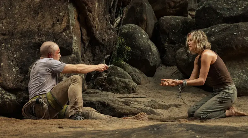 Charlize Theron atada a un árbol mientras Taron Egerton se agacha a su lado con una cesta.