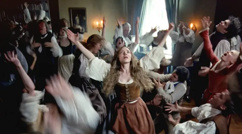 Fotograma De 'El Testamento De Ann Lee'. Amanda Seyfried En El Centro De Una Habitación Rodeada De Personas Moviéndose Frenéticamente Con Los Brazos En Alto.