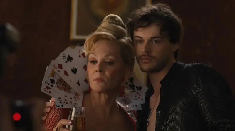 Fotograma De La Temporada 5 De 'Hacks'. Deborah Vance Luciendo Un Cuello Hecho De Naipes De Póker Junto A Un Hombre Con Camisa Negra.