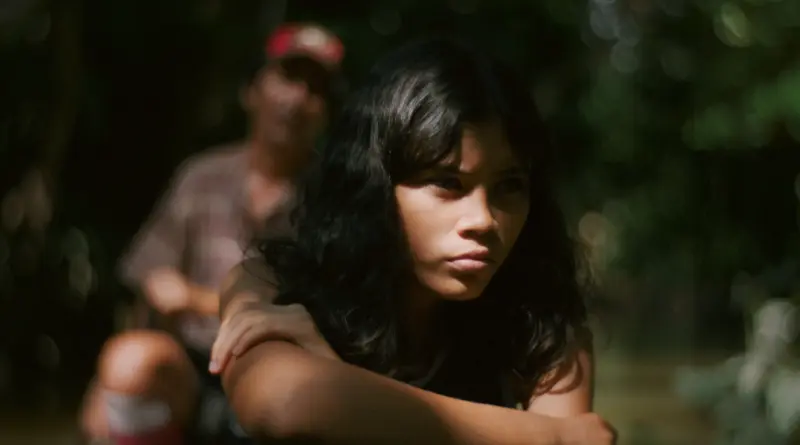 Fotograma De 'Manas'. La Protagonista Sentada En Un Bosque Con Los Brazos Cruzados Y Mirada Seria, Con Una Figura Borrosa Al Fondo.