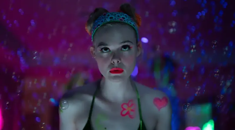 Fotograma De La Serie 'Margo Tiene Problemas De Dinero'. Elle Fanning Con Maquillaje Colorido Y Una Diadema Azul, Rodeada De Burbujas Flotantes Bajo Una Iluminación De Neón Rosa Y Azul.