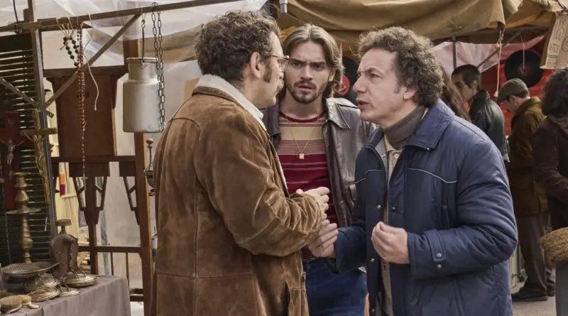 Fotograma De 'Por Cien Millones'. Tres Hombres Con Atuendos Retro De Los Años 70 Conversando De Forma Tensa En Medio De Un Mercado Callejero.