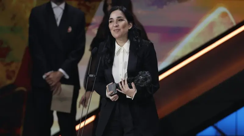 Imagen De Los Premios Goya 2026. Una Mujer, Alauda Ruiz De Azúa, Vestida Con Traje Negro Y Camisa Blanca Sostiene Un Premio Goya Y Sonríe Frente A Un Micrófono En El Escenario De La Gala.