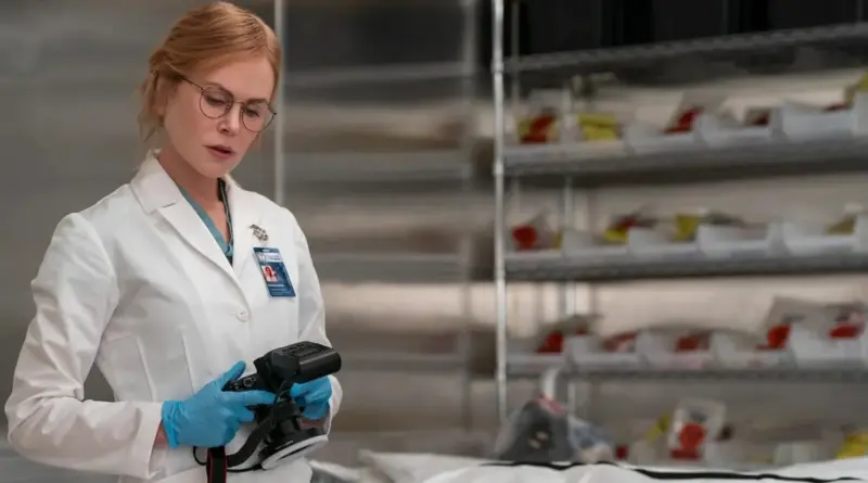 Fotograma De 'Scarpetta'. Nicole Kidman Caracterizada Como Doctora Forense Con Bata Blanca Y Gafas, Revisando Una Cámara Fotográfica Profesional En El Laboratorio.