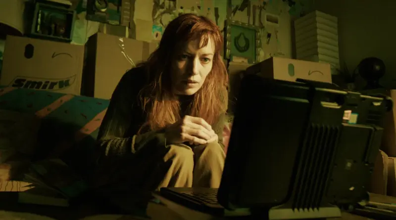 Fotograma De 'Sender'. Britt Lower, Con Pelo Rojizo, Mira Fijamente La Pantalla De Un Ordenador En Una Habitación Llena De Cajas Smirk.