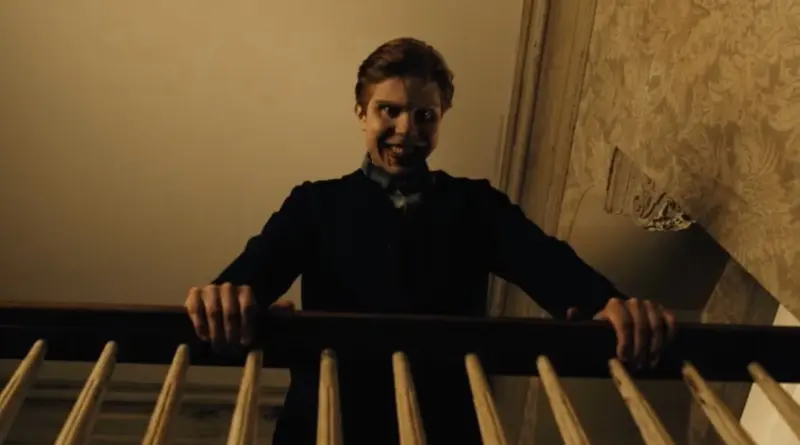 Primer plano en ángulo bajo de un hombre con una sonrisa siniestra y exagerada mirando desde una barandilla de madera en la serie de terror "From".