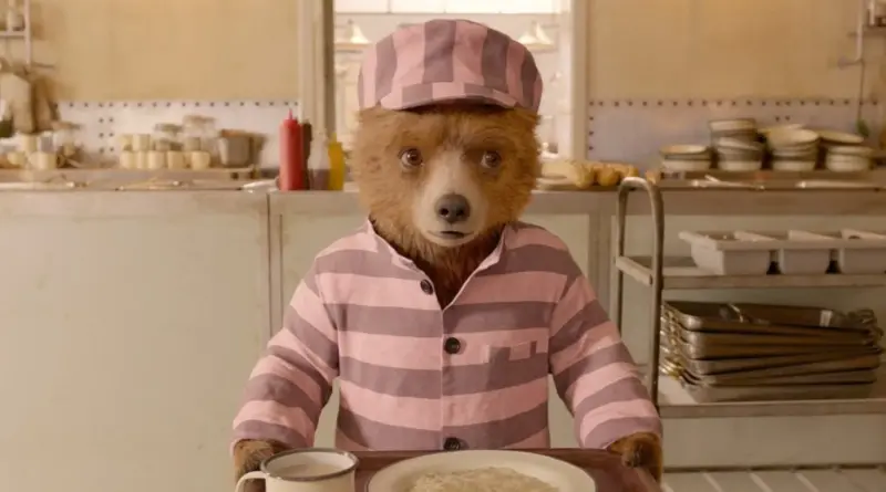 El oso Paddington con uniforme de preso a rayas rosas y blancas sosteniendo una bandeja de comida.