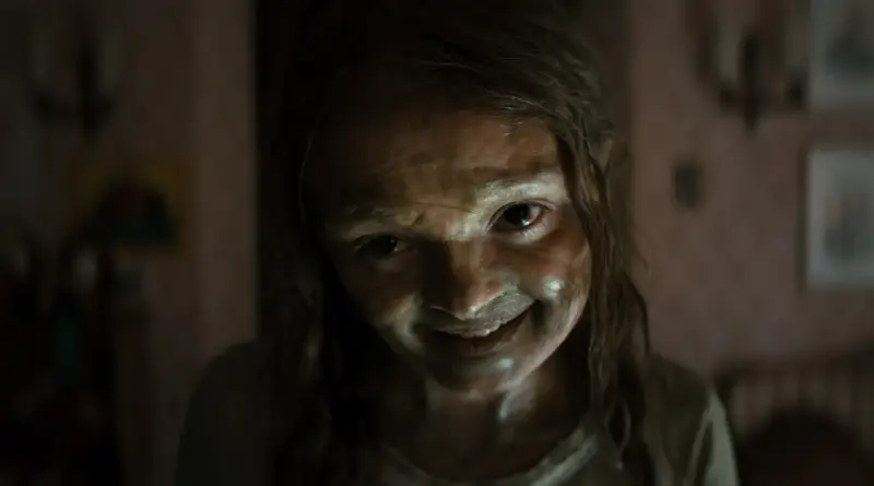 Personaje con una sonrisa siniestra y rostro pálido en una escena oscura de La momia de Lee Cronin.