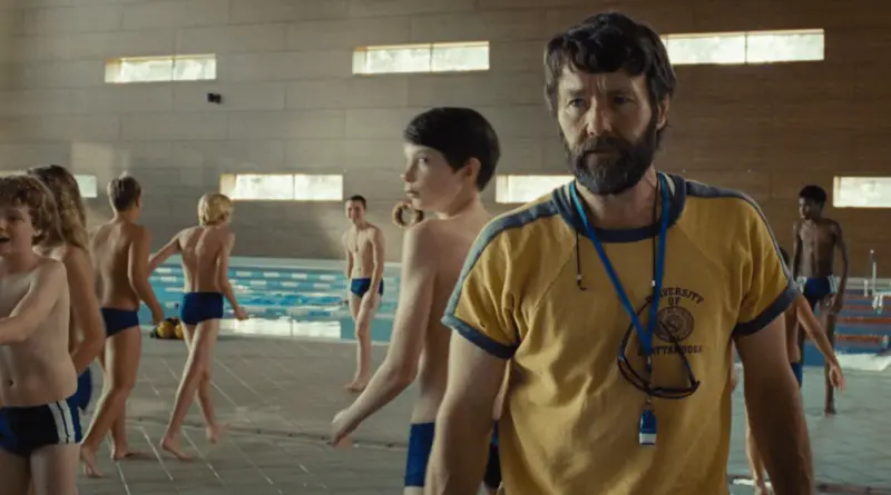 Un entrenador con barba en primer plano y niños nadadores al fondo en una piscina cubierta de la película La Plaga.
