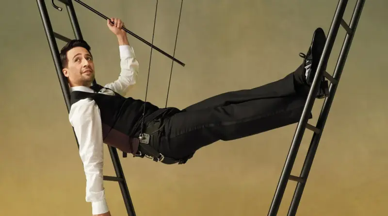 Lin-Manuel Miranda, director de 'Octet', suspendido en el aire con arneses y cables, posando de forma horizontal entre dos escaleras negras sobre un fondo degradado cálido.