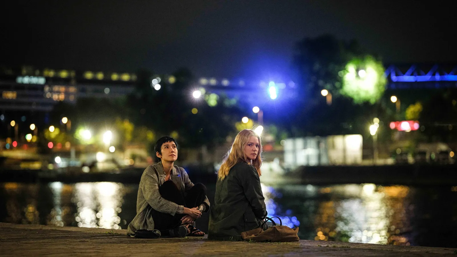 Fotograma de la película All of a Sudden (2026) donde dos mujeres están sentadas al borde de un río por la noche con las luces de la ciudad reflejadas en el agua.