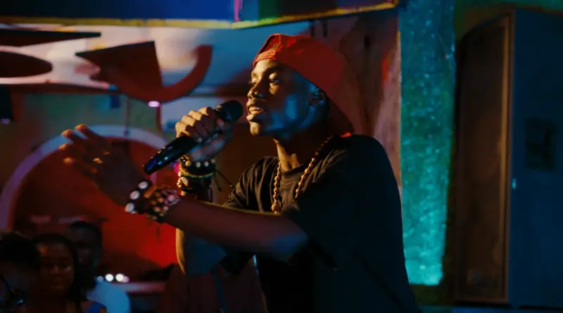 Retrato dinámico y lleno de energía de un joven intérprete en la película "Congo Boy", capturado a mitad de la actuación en el escenario con un micrófono en la mano y una expresión apasionada bajo iluminación teatral de neón azul y rojo.